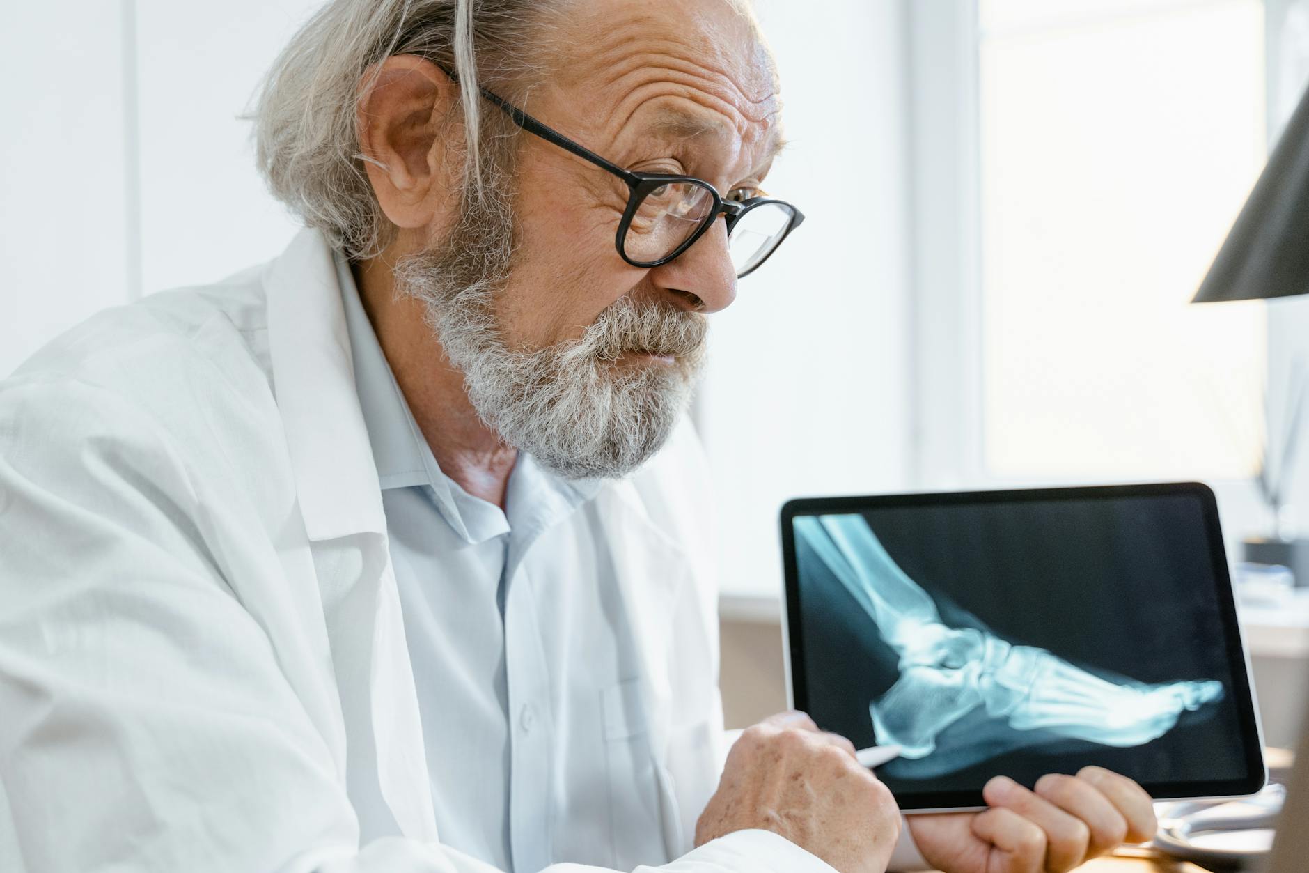 Elderly orthopedic doctor in white coat examining a foot X-ray on a digital tablet.